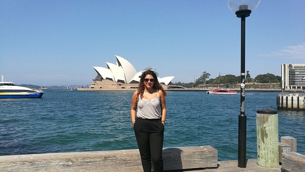 chica morena con camiseta de tirantes, pantalón negro y gafas de sol, posando y detrás la Ópera House de Sydney